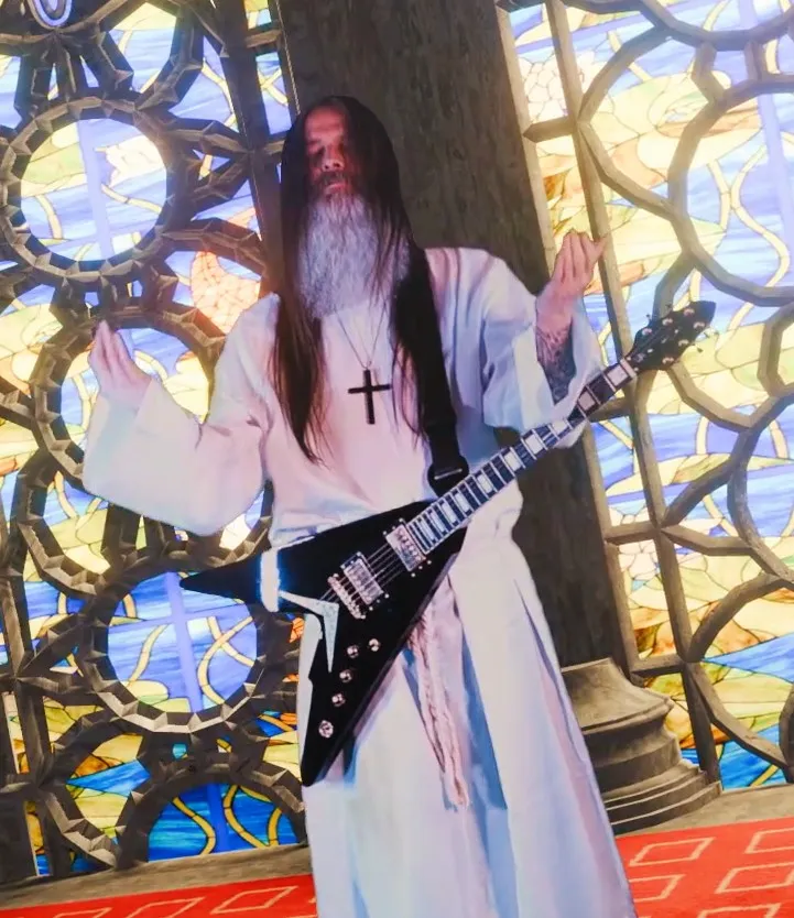 Peter Capon in a white robe with a flying-V guitar in front of stained glass